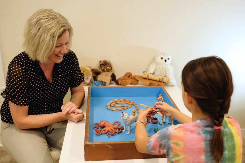 Kristina laughs, hands folded on the table, as a child seen from behind sets a wolf, a giraffe and a winged dragon into the sand tray.