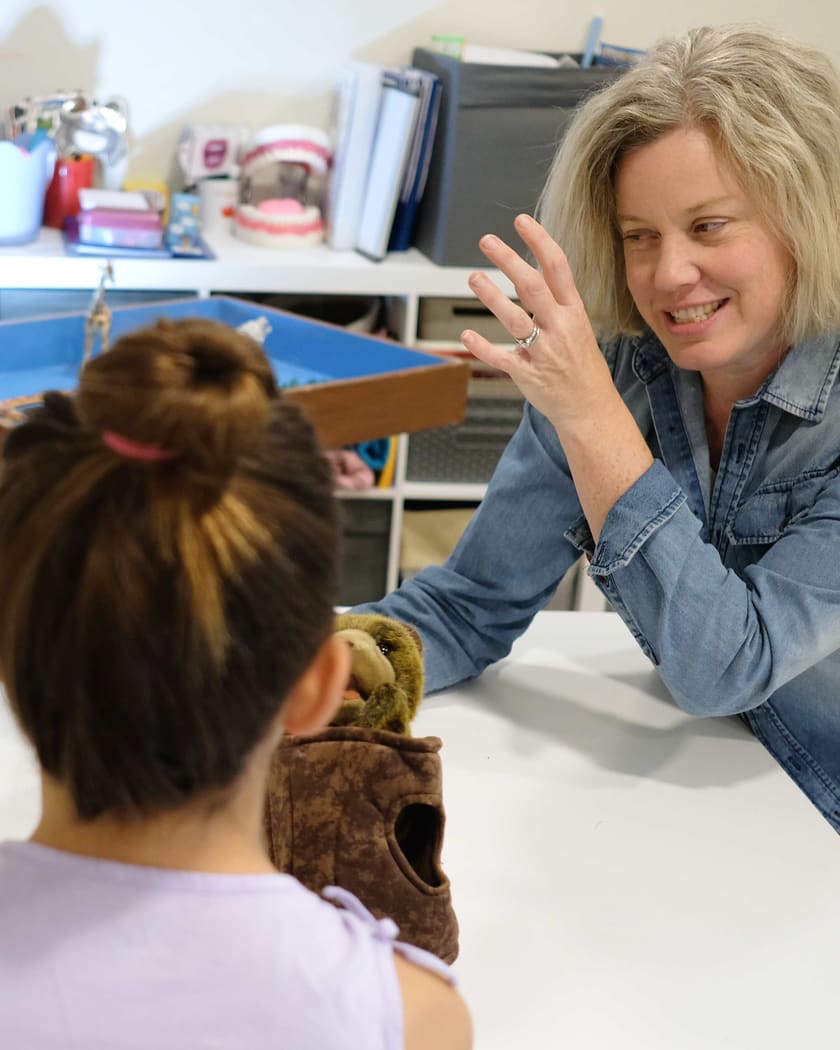 A child, seen from behind, holds a puppet up across the play-room table to Kristina, who smiles back with one hand raised. The sand tray sits on the shelf behind them.